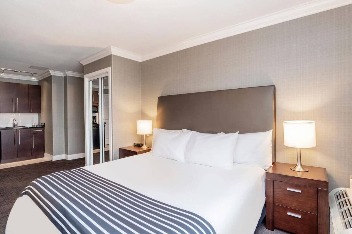 A hotel room with a large bed, two bedside tables with lamps, a separate bathroom, and a kitchenette.
