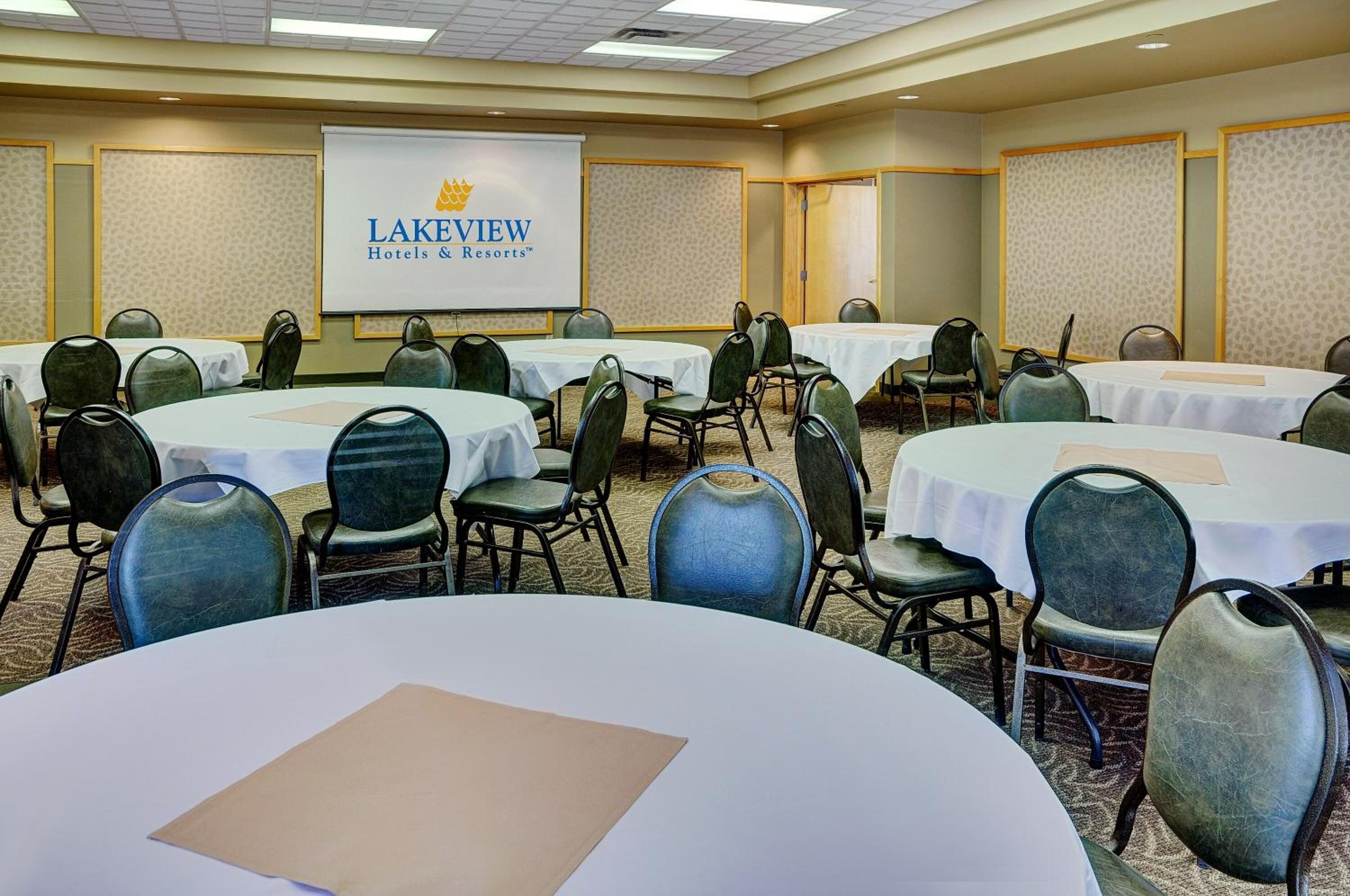 Una sala conferenze con tavoli e sedie rotonde, uno schermo proiettore con il logo di Lakeview Hotels & Resorts e un pavimento rivestito di moquette.