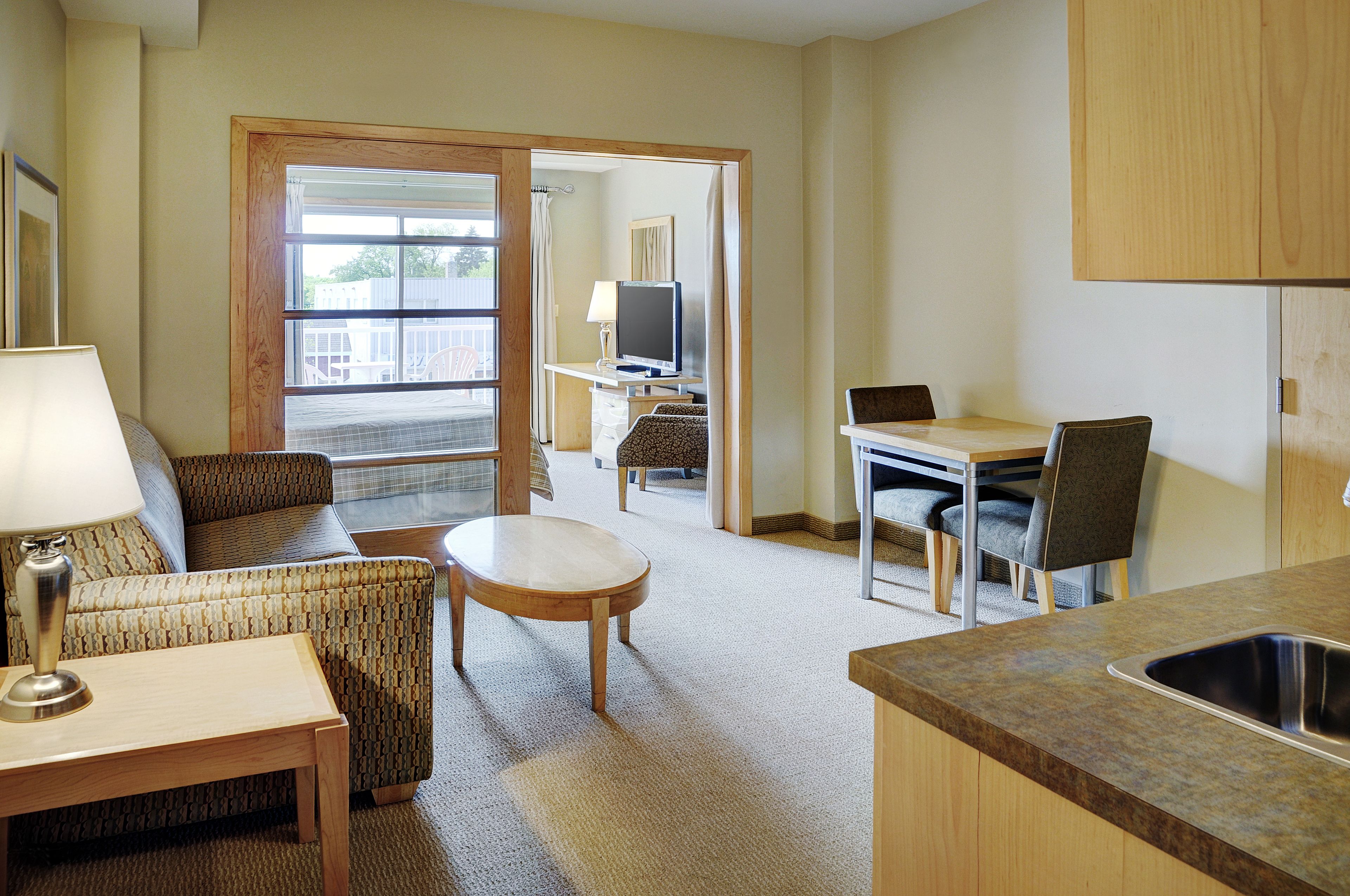 A compact hotel room with a kitchenette, a dining area, a seating area with a sofa and armchair, and a television.