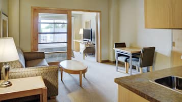 A compact hotel room with a kitchenette, a dining area, a seating area with a sofa and armchair, and a television.