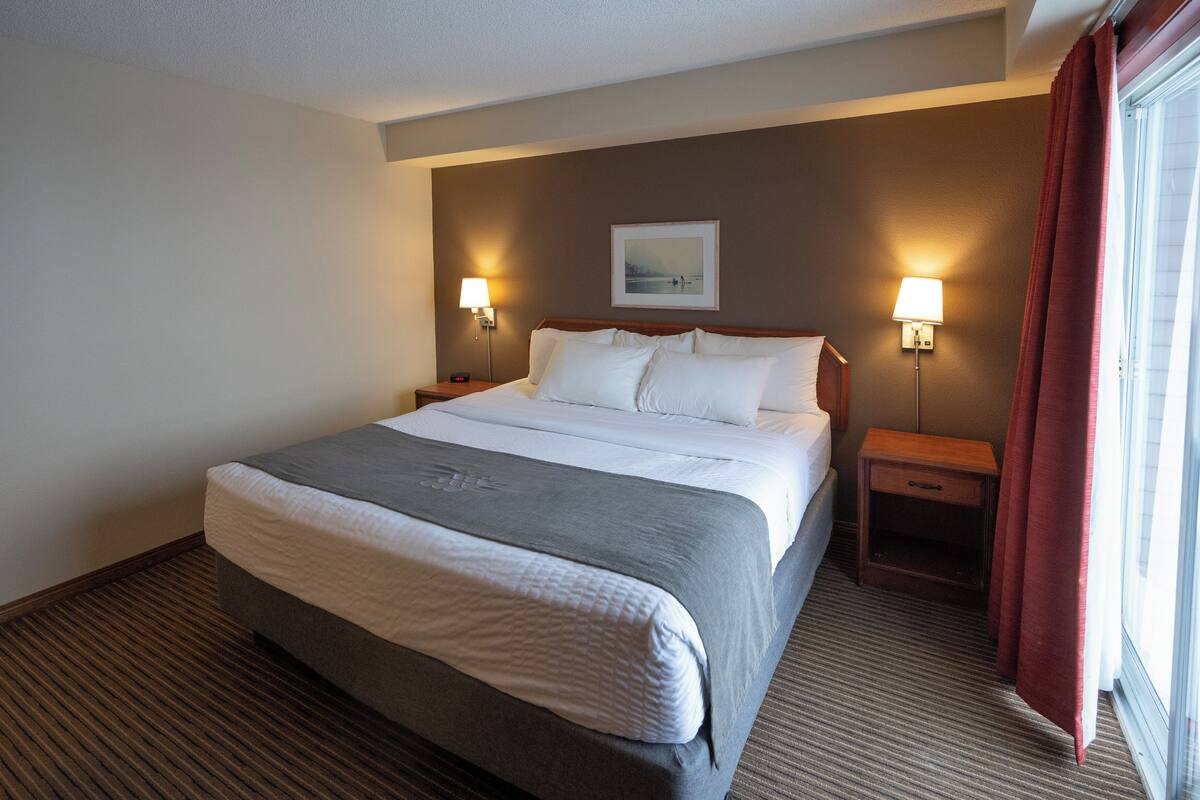 Habitación de hotel con una cama grande, dos mesitas de noche con lámparas, un cuadro enmarcado en la pared y una ventana con cortinas.