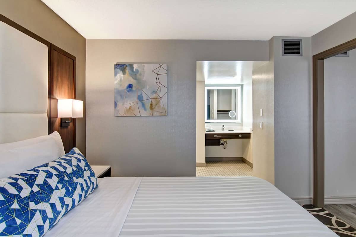 A hotel room with a bed, a nightstand, a lamp, a painting, a bathroom with a sink and mirror.