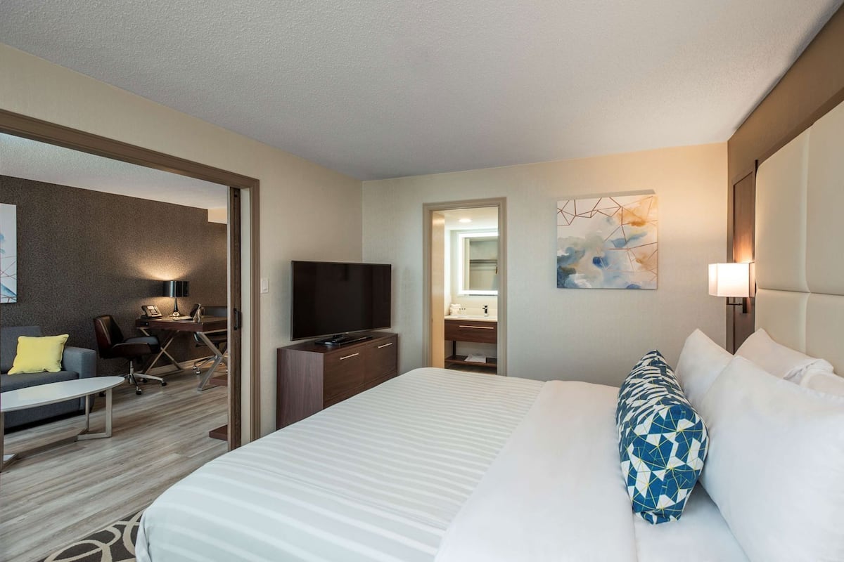 A hotel room with a bed, a desk with a TV, a chair, a sofa, and a bathroom visible through an open door.