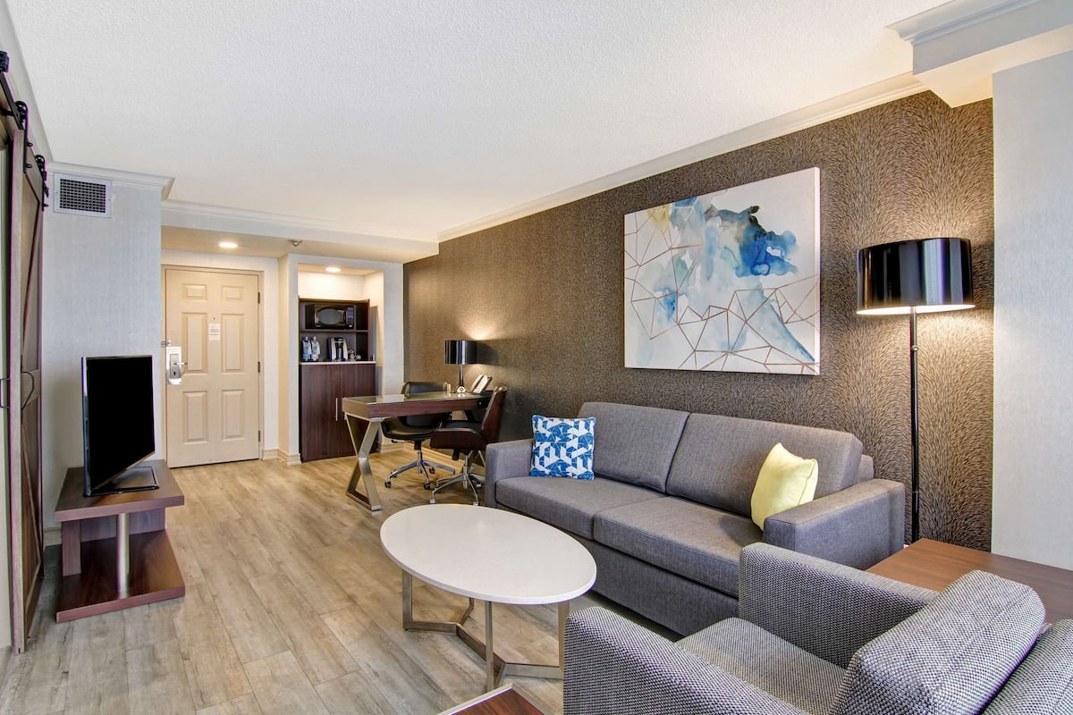 A modern living room with a grey sofa, a white coffee table, and a flat-screen TV on a wooden stand. There is a dining area with a table and chairs, and a kitchen in the background.