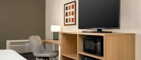 Habitación de hotel con cama, escritorio con lámpara, silla, televisor, microondas y lavavajillas.