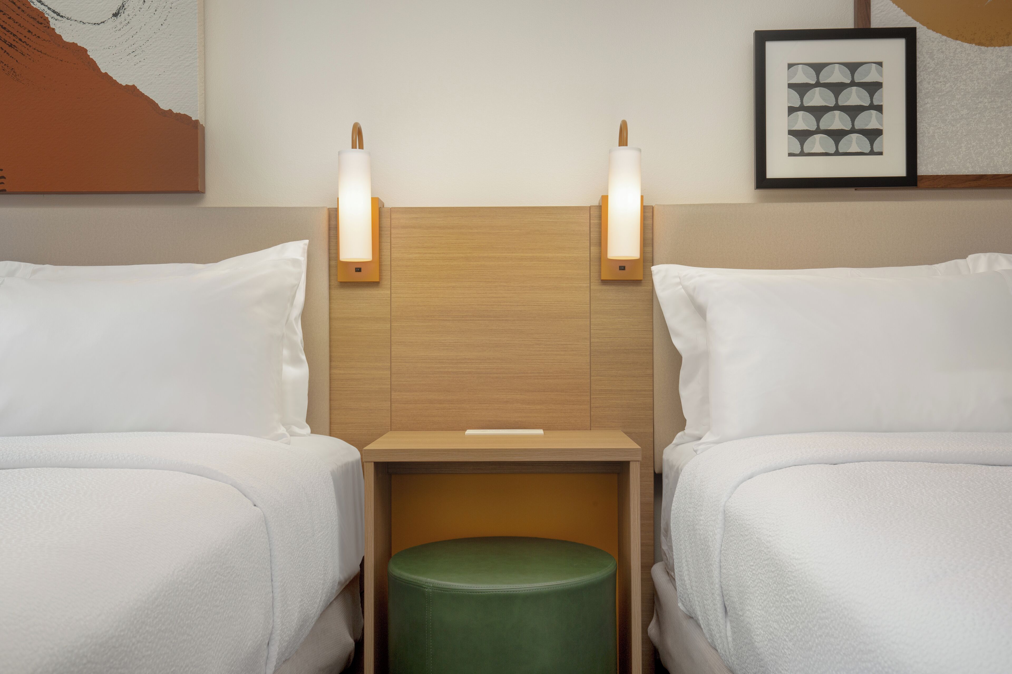 Een hotelkamer met twee bedden, een houten hoofdbord, bedlampjes, een groene poef en een lijst met een kunstwerk aan de muur.