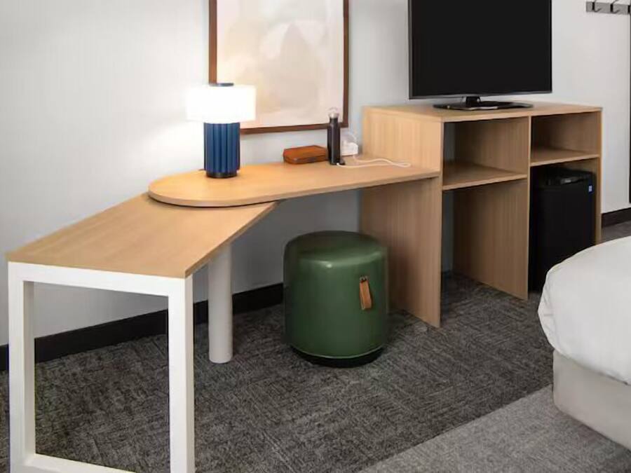 A hotel room with a wooden desk, a green trash bin, a bed, and a TV on a stand.