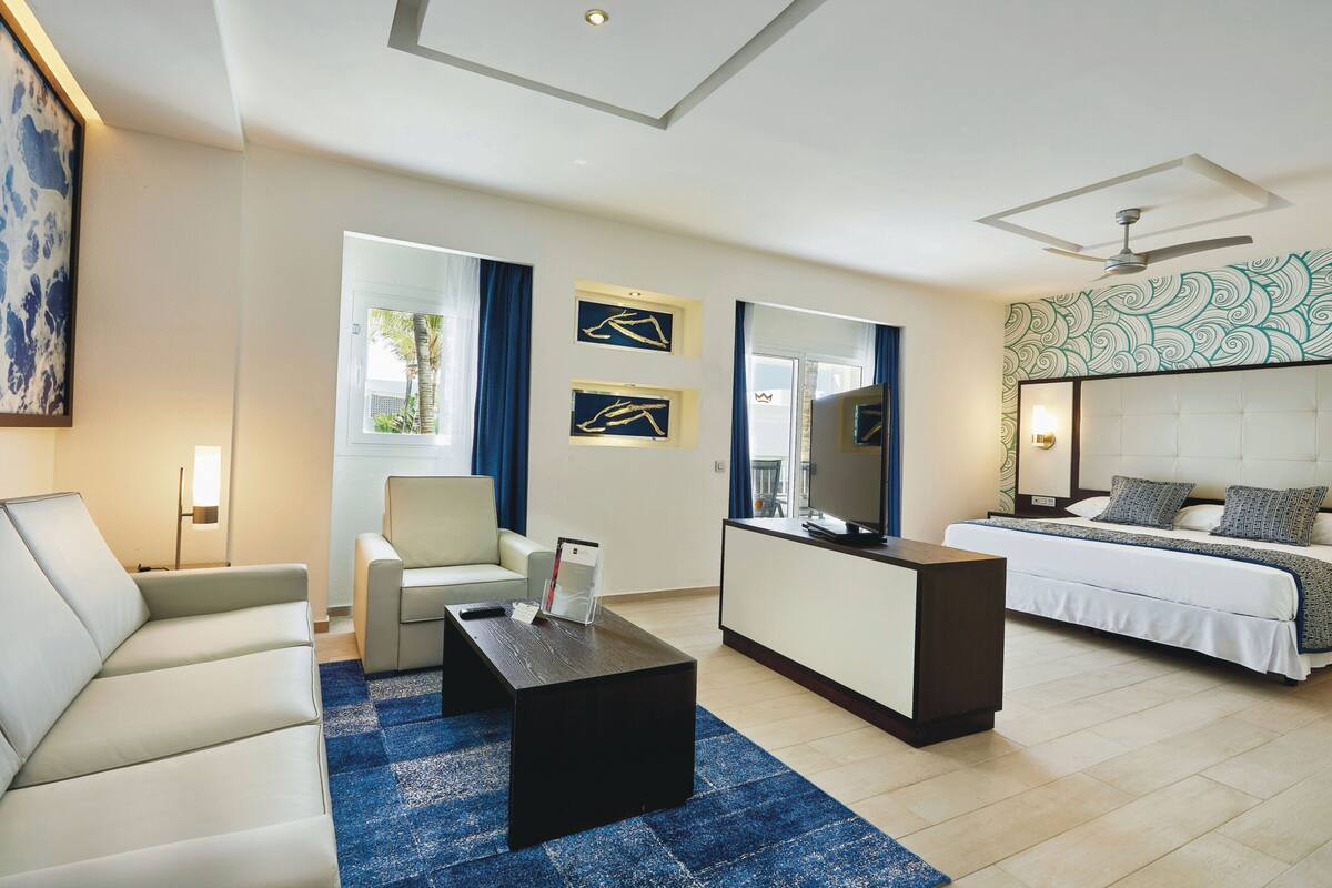 A modern hotel room with a sofa, a coffee table, a TV, a bed with a headboard, and artwork on the walls.