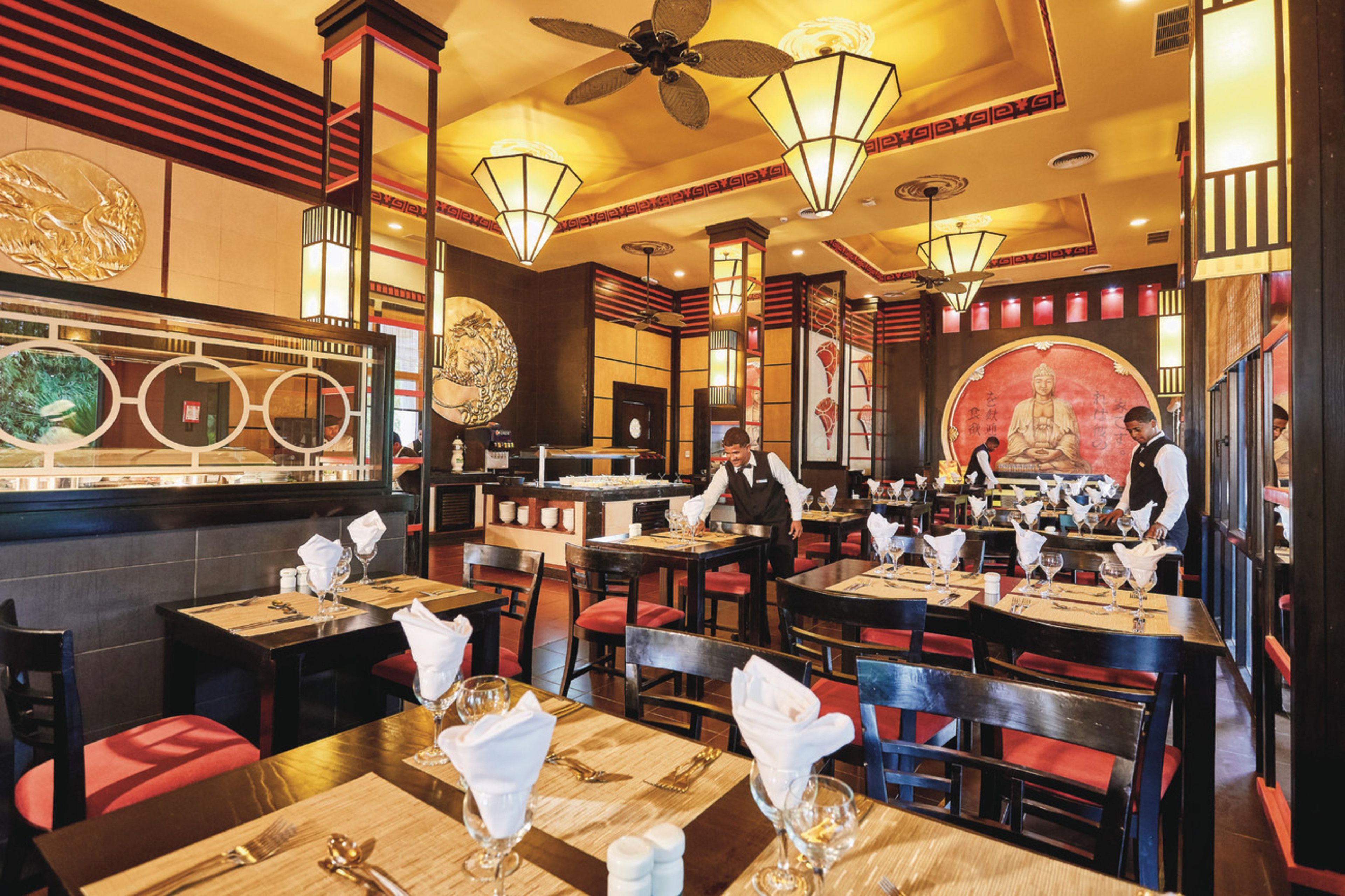 Un restaurante con varias mesas, sillas rojas acolchadas y una estatua de Buda al fondo.