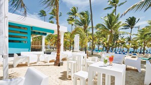 A beachside restaurant with white tables and chairs, palm trees, and a view of the ocean.