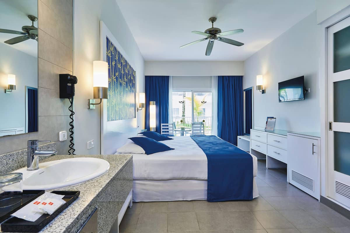 A hotel room with a large bed, a bathroom with a sink and mirror, a TV, and a ceiling fan.
