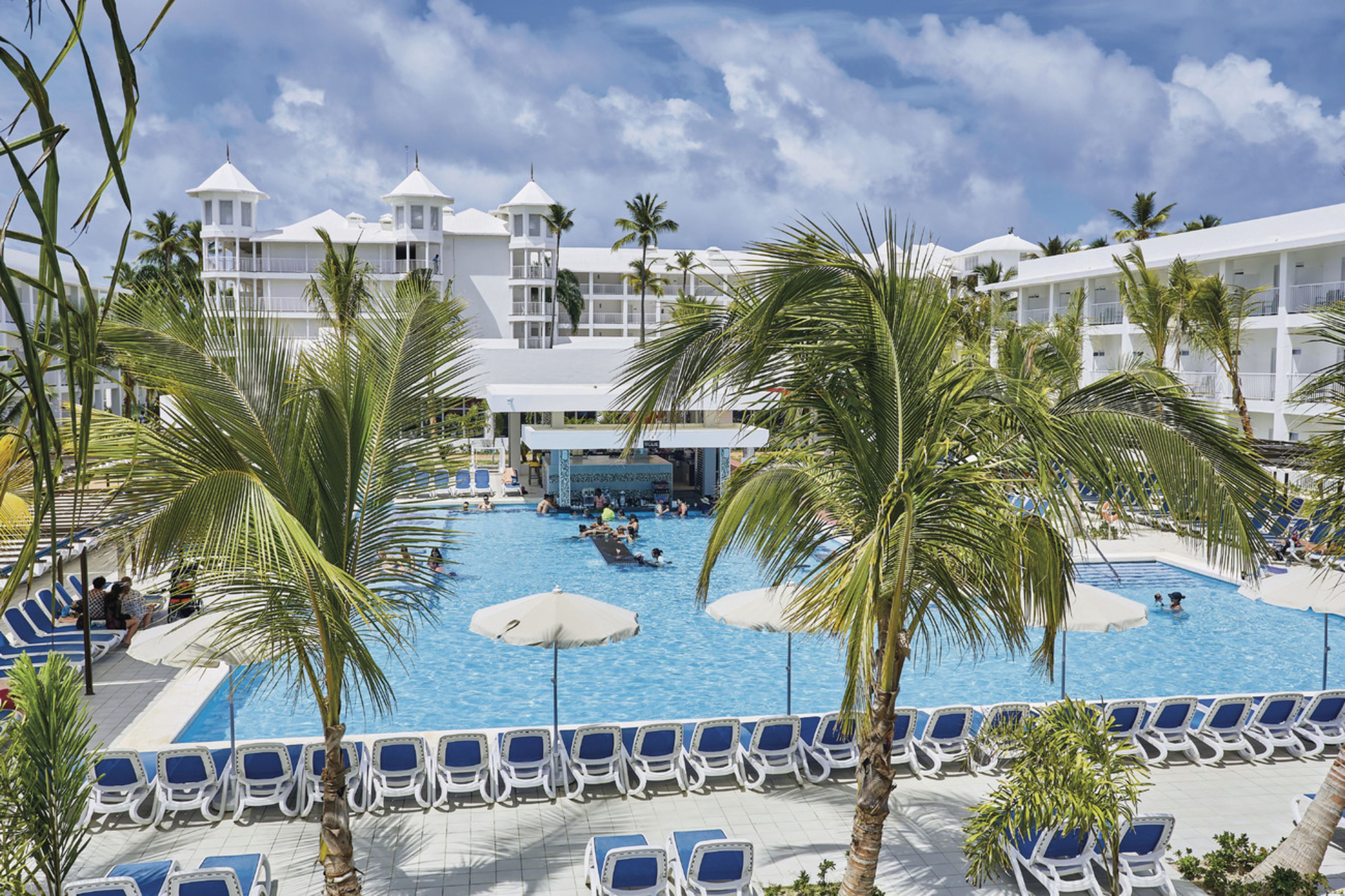 Un área de piscina de hotel con múltiples sillones de descanso, palmeras y una gran piscina.