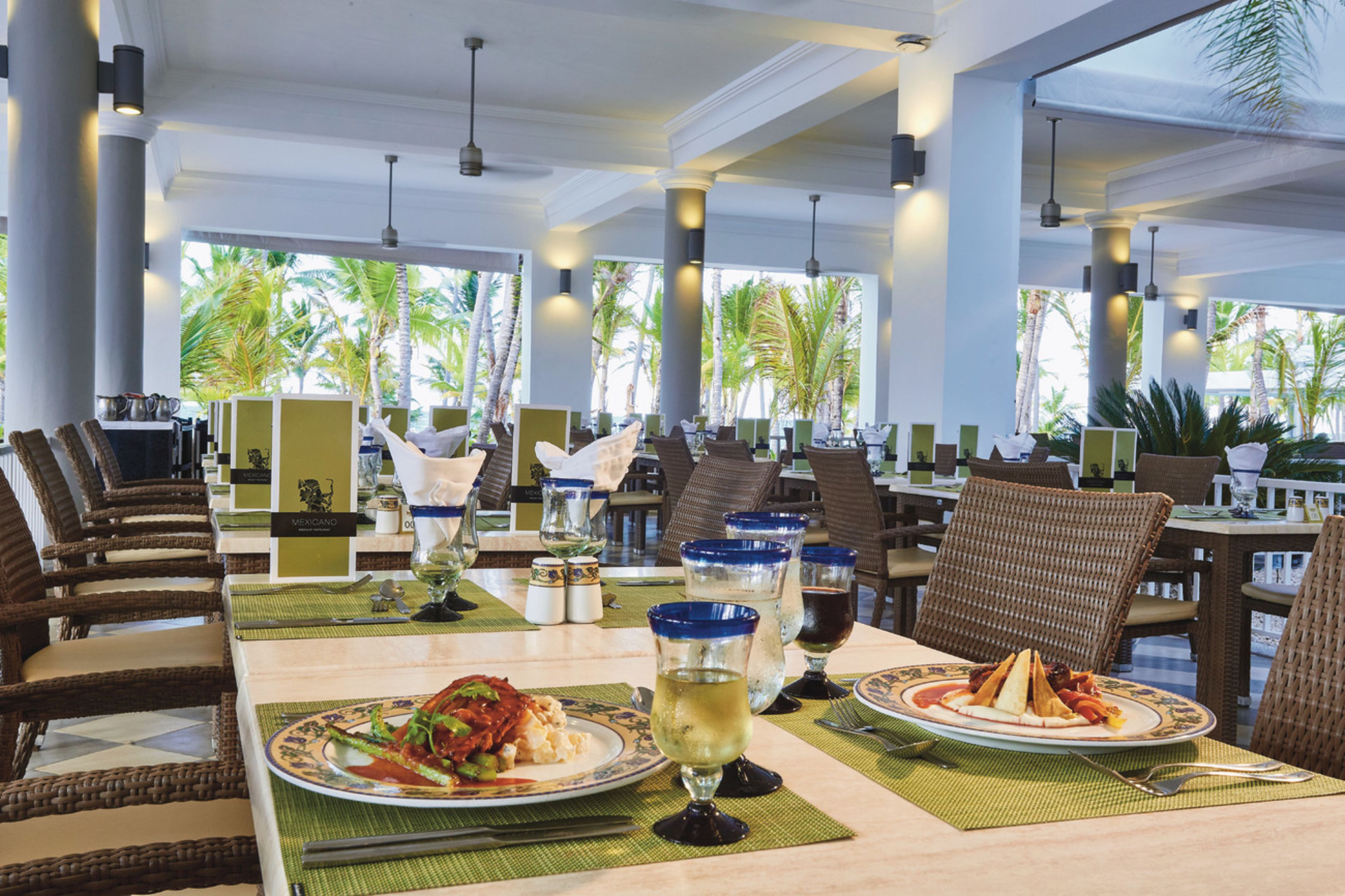 A restaurant with tables set for dining, featuring a view of palm trees outside.