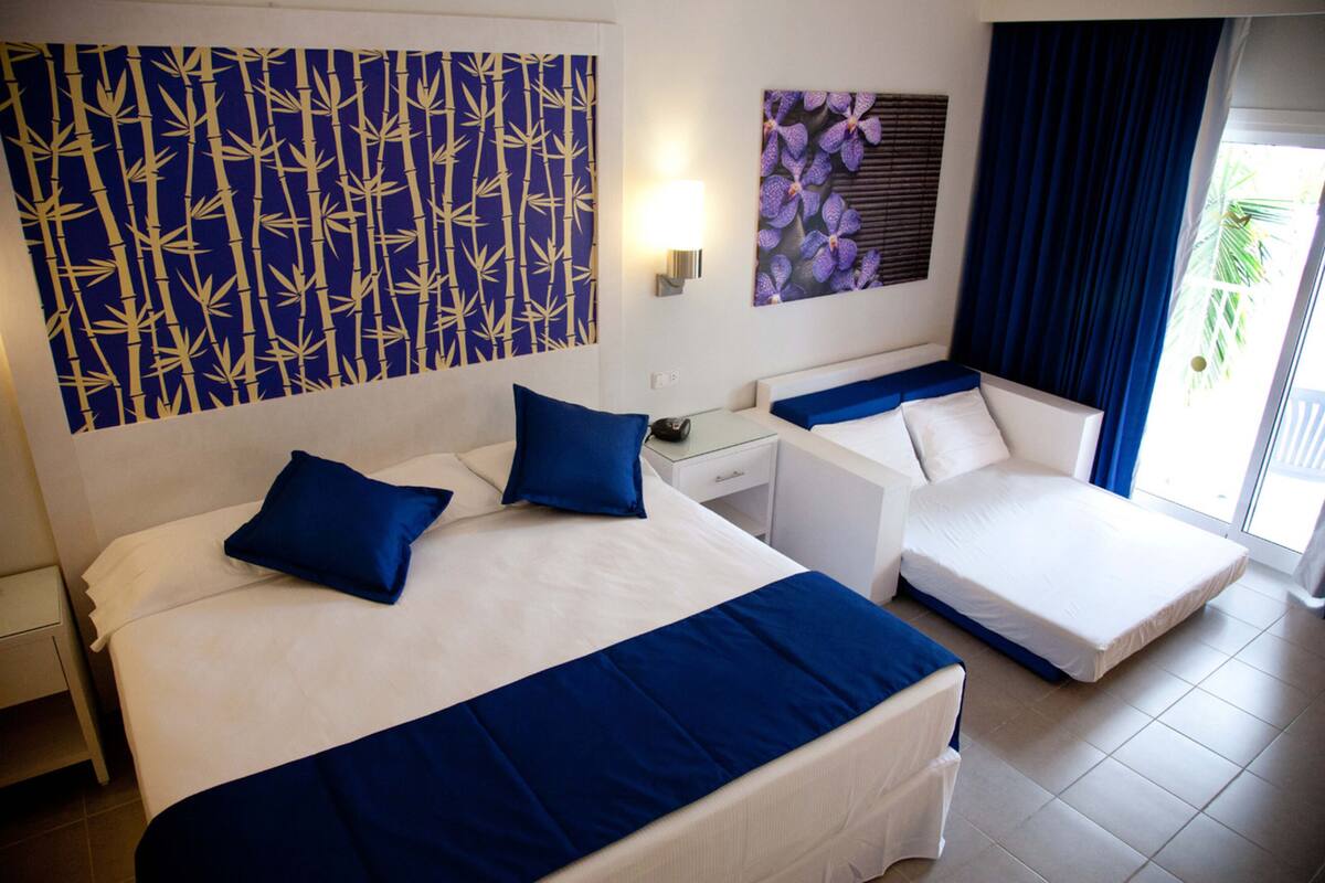 A hotel room with two beds, a nightstand, a lamp, and a window with curtains.