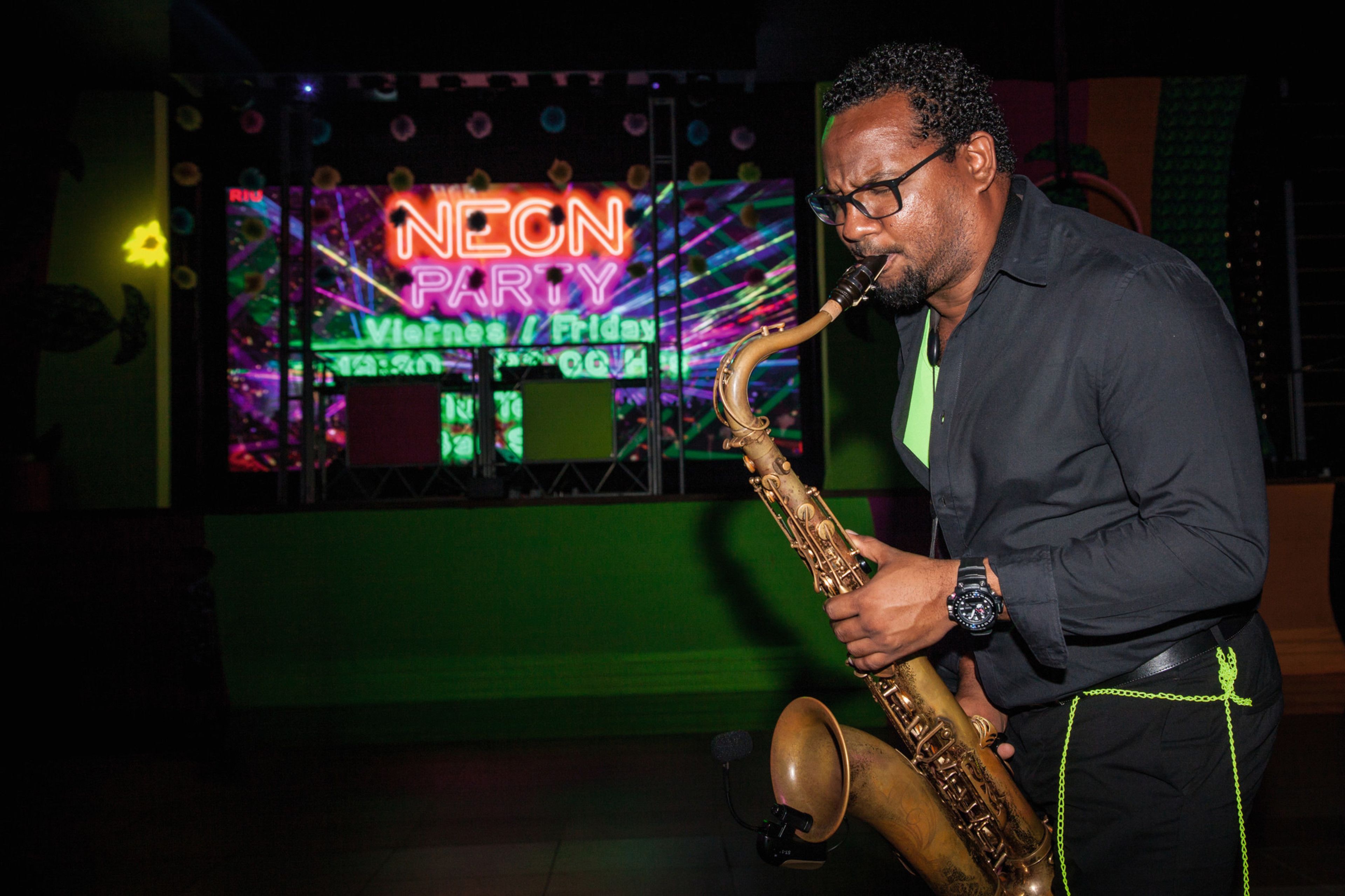 Una persona tocando el saxofón en un local iluminado con neón, con un cartel que dice "NEON PARTY".