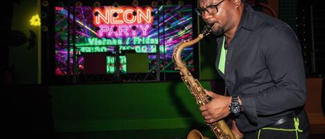 Una persona tocando el saxofón en un local iluminado con neón, con un cartel que dice "NEON PARTY".