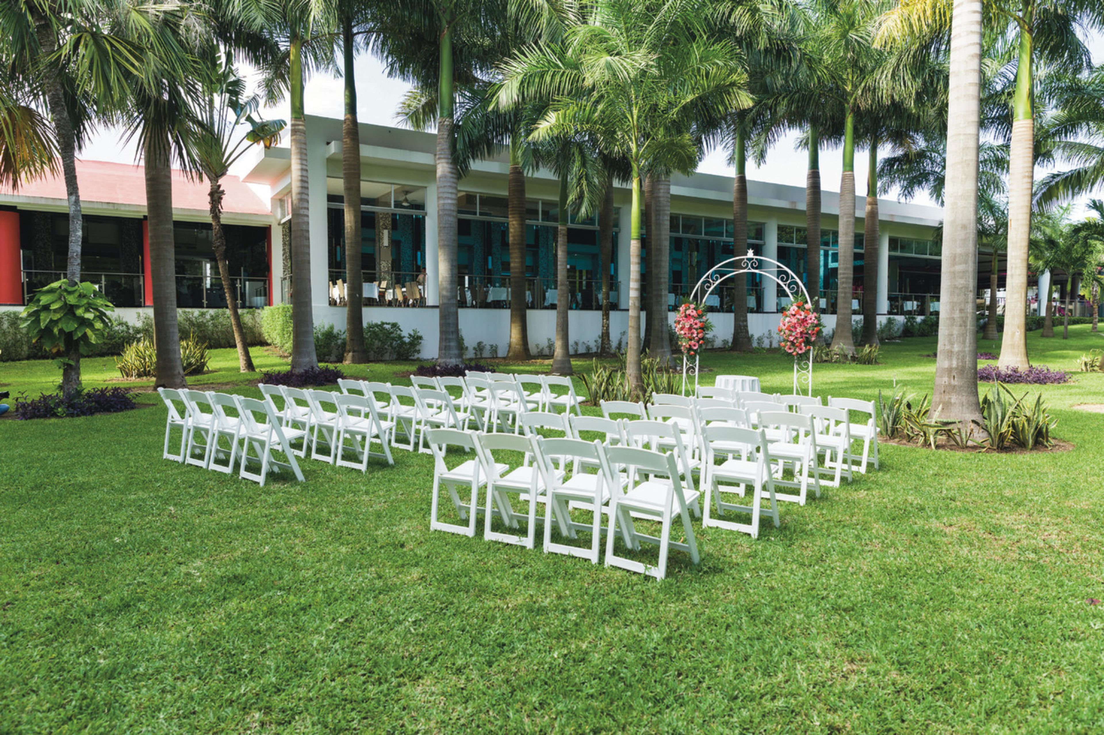 Une cérémonie de mariage en plein air, avec des chaises blanches disposées en rangées, une arche décorée de fleurs et des palmiers.