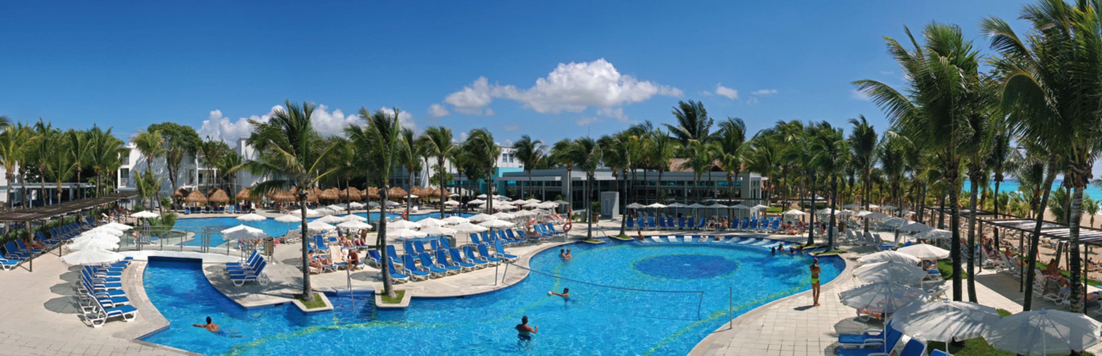 Una amplia piscina al aire libre con varias pistas de natación, rodeada de tumbonas y sombrillas, y palmeras al fondo.
