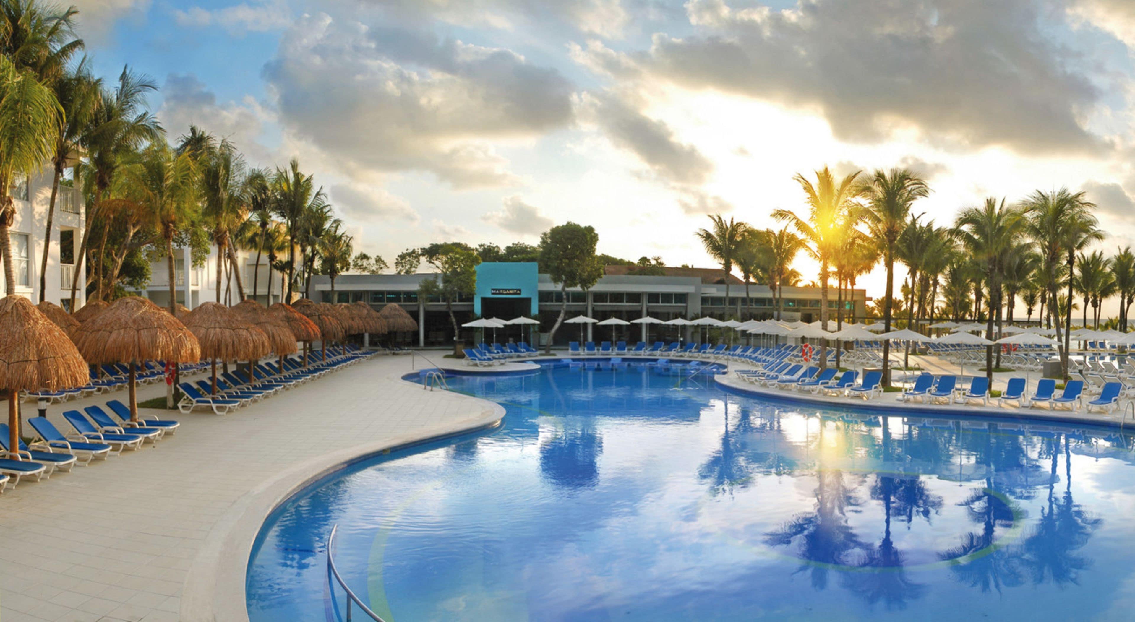 A resort pool area with blue lounge chairs and thatched umbrellas, surrounded by palm trees and a building labeled "MARGARITA".