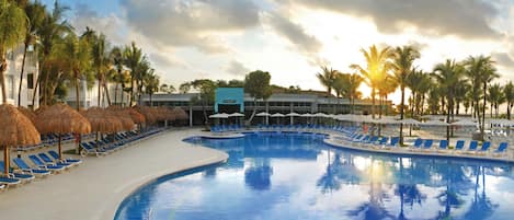 A resort pool area with blue lounge chairs and thatched umbrellas, surrounded by palm trees and a building labeled "MARGARITA".