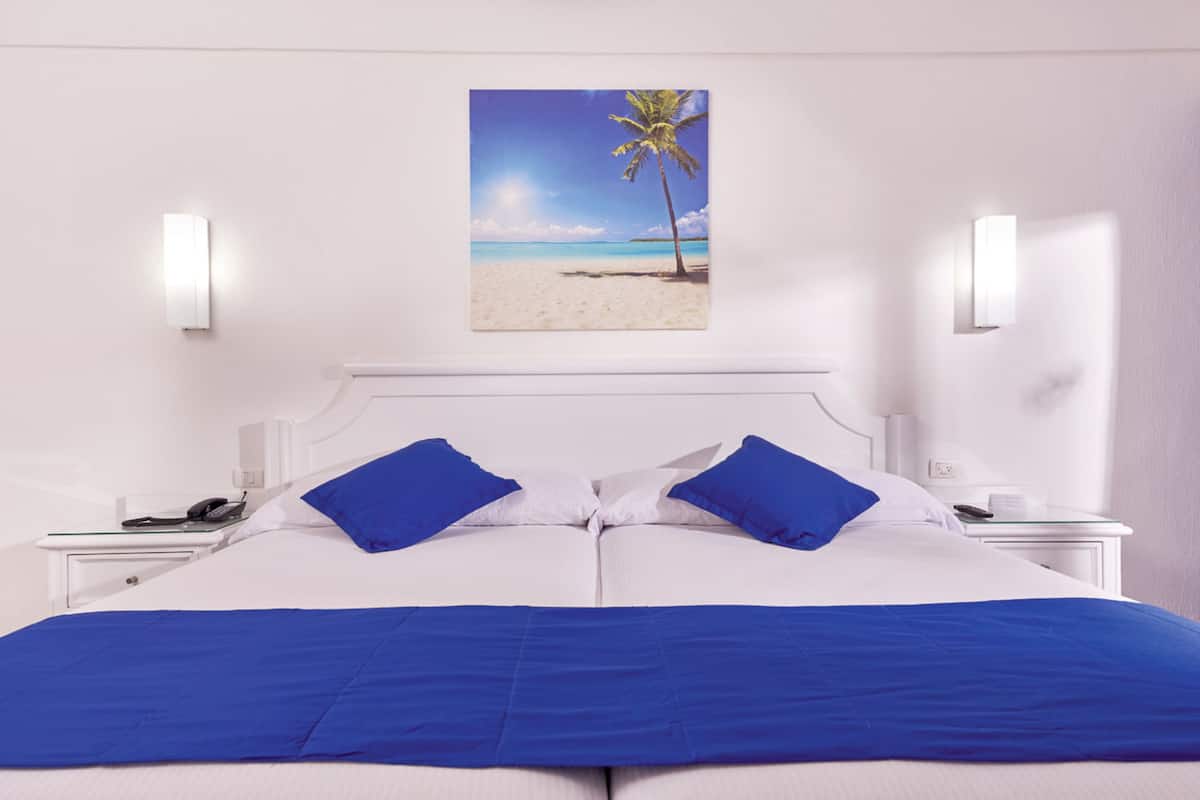A hotel room with a bed, two blue pillows, a white headboard, a nightstand with a phone, and a wall-mounted picture of a beach.