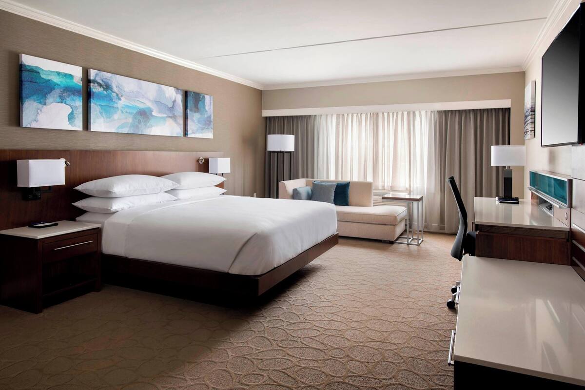 Habitación de hotel con una cama grande, un escritorio, una silla y un televisor. Hay dos cuadros en la pared y una ventana con cortinas.