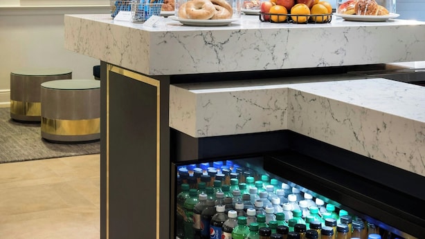 A hotel breakfast bar with a marble countertop, a hanging light fixture, and a refrigerated beverage dispenser below. - Delta Hotels by Marriott Basking Ridge (Basking Ridge)