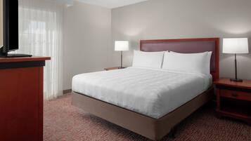 A hotel room with a large bed, two bedside tables with lamps, a TV on a stand, and a window with sheer curtains.