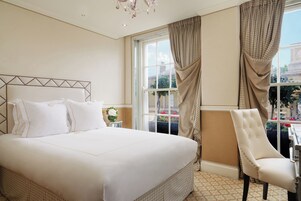 A hotel room with a large bed, a chair, a window with curtains, and a chandelier.