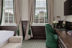 Hotelkamer met een bed, een groene fauteuil, een houten dressoir, een flatscreen televisie en uitzicht op gebouwen door grote ramen met gordijnen.