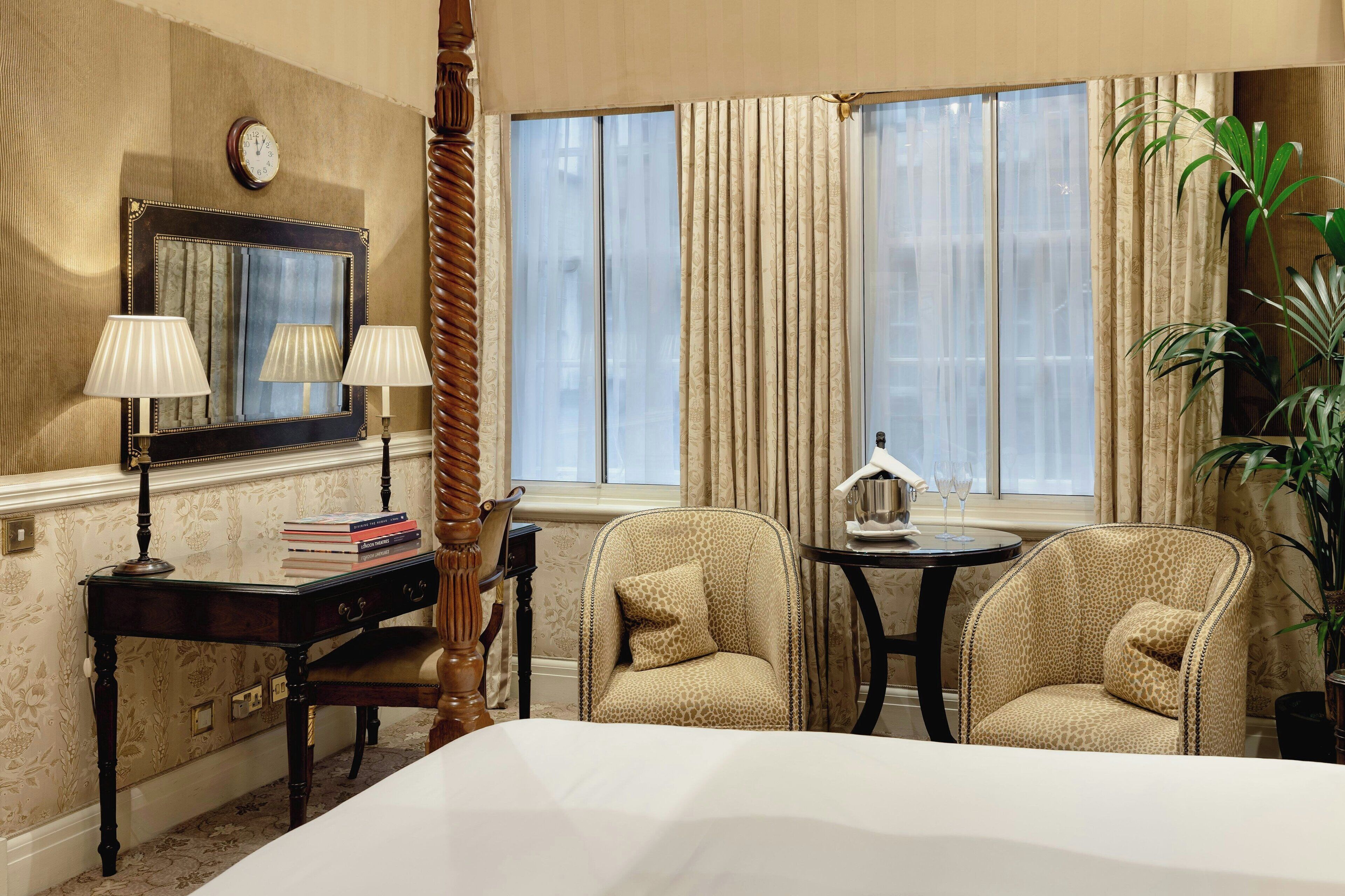 Ein Hotelzimmer mit einem Bett, zwei Sesseln, einem kleinen Tisch mit Lampe, einem Spiegel, einer Uhr und einem Fenster mit Vorhängen.