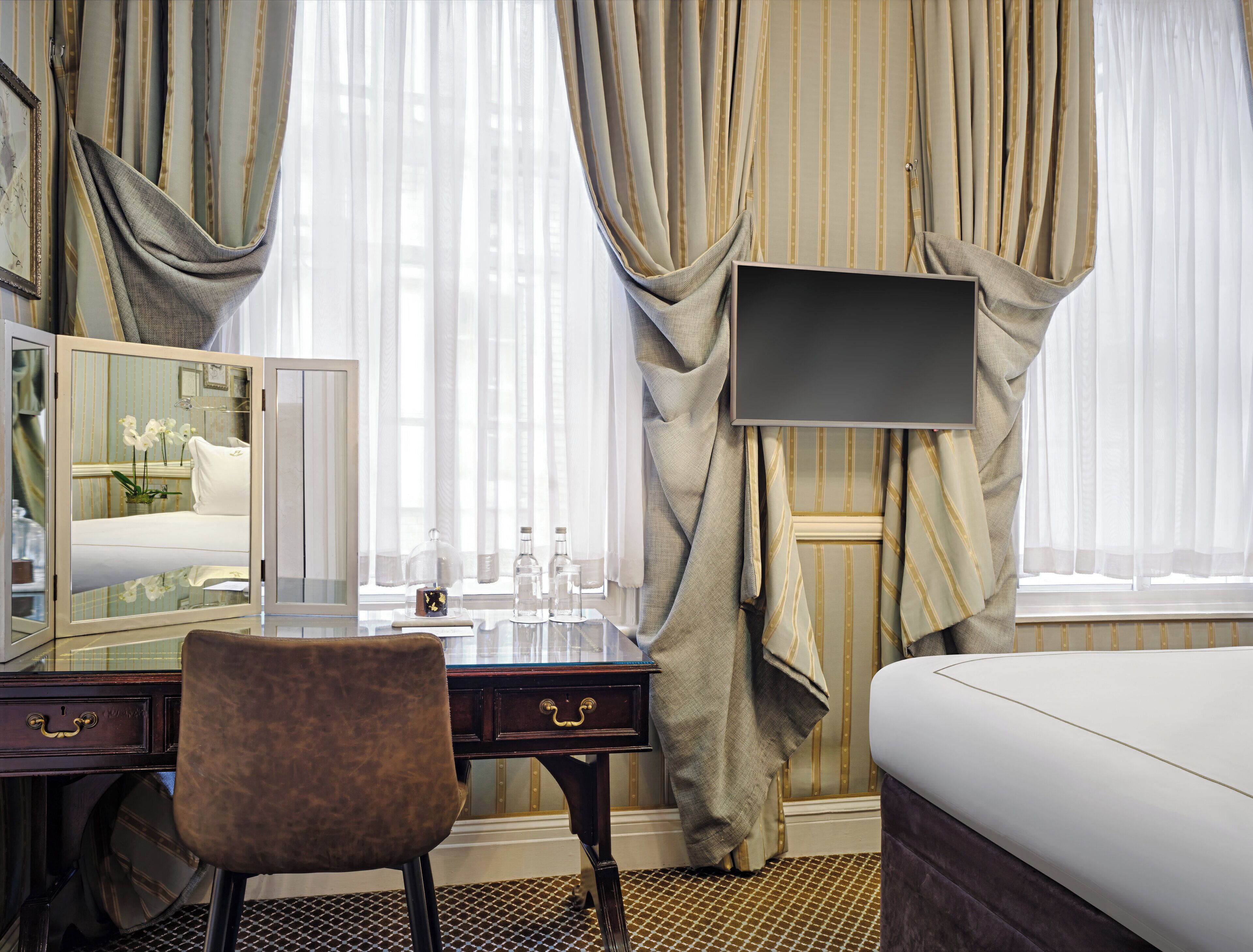 A hotel room with a bed, a desk with a mirror, a chair, and a TV mounted on the wall.