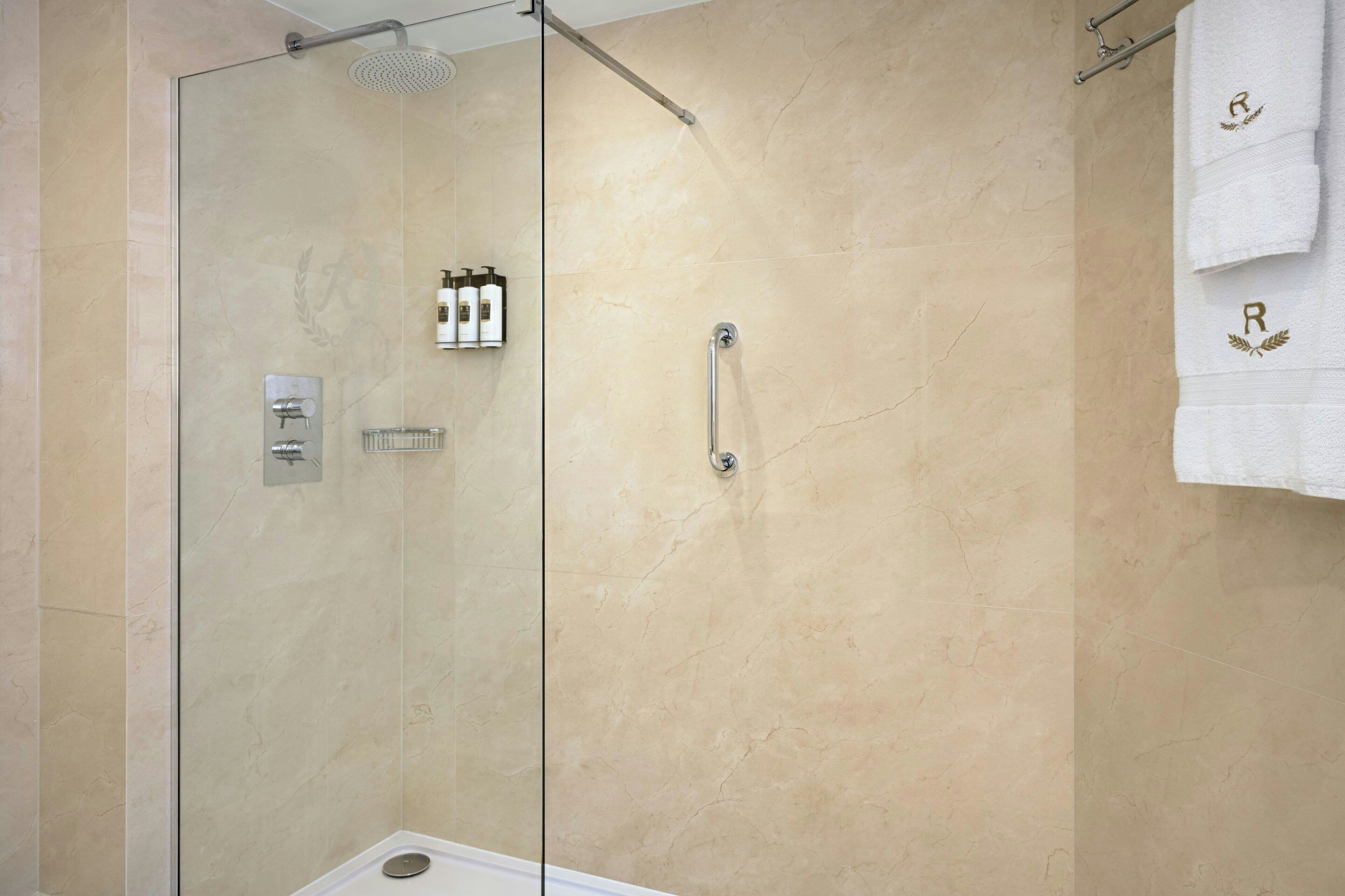 A glass-enclosed shower with a handheld showerhead, a towel rack with towels, and a wall-mounted soap dispenser.