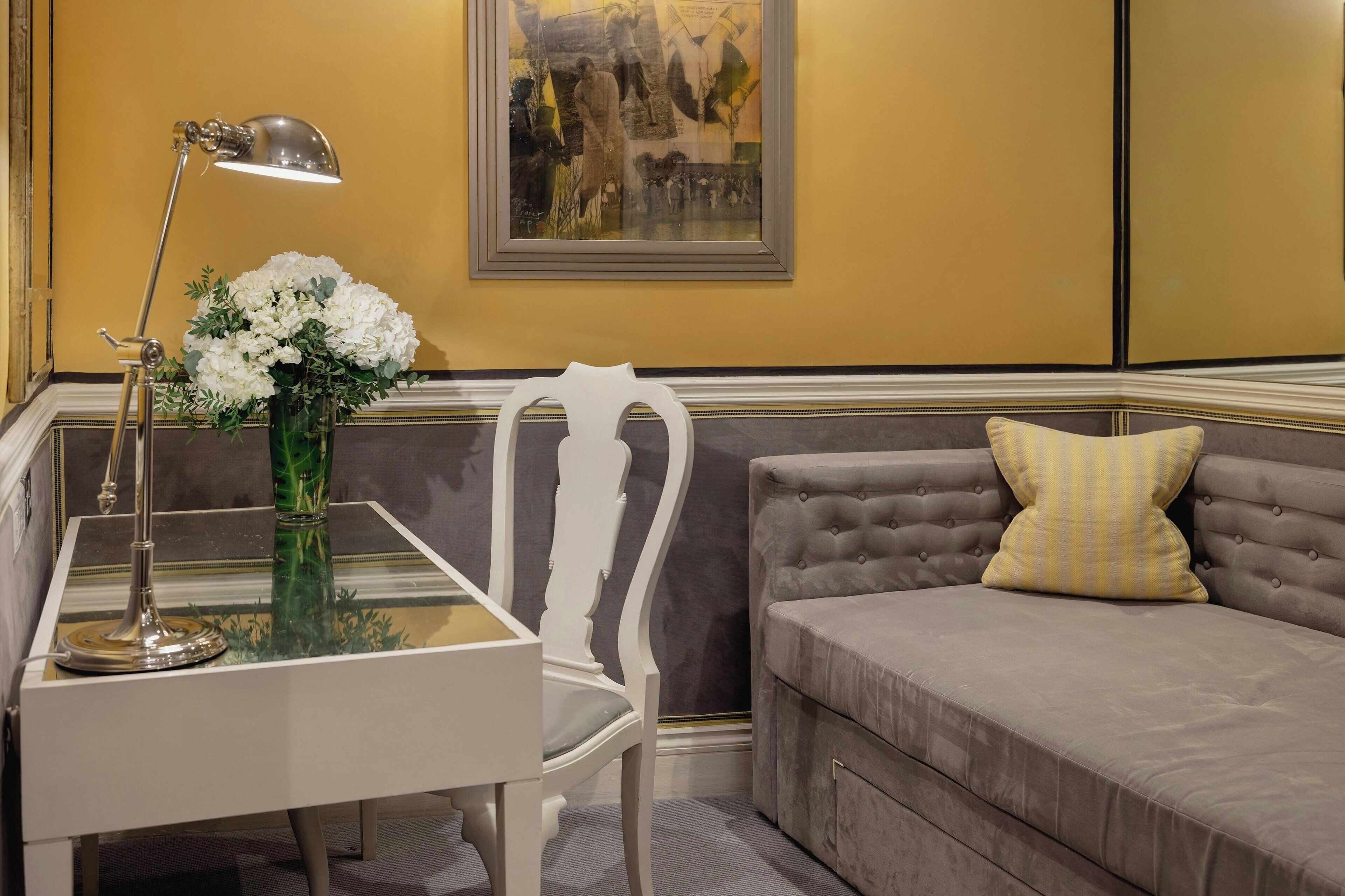 A room with a glass-top table, a white chair, a grey sofa with a yellow pillow, and a framed abstract painting on the wall.