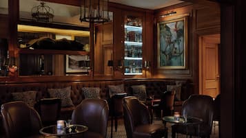 A dimly lit bar area with leather seating, a bar counter with bottles, and a large framed painting on the wall.