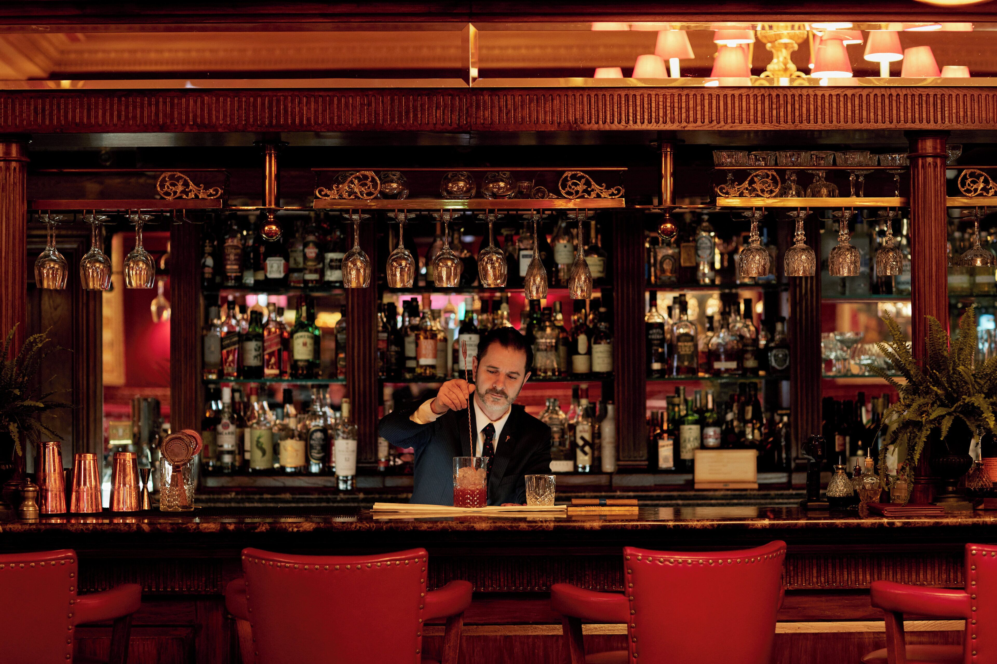 A bar area with a bartender, red bar stools, and shelves stocked with bottles and glasses.
