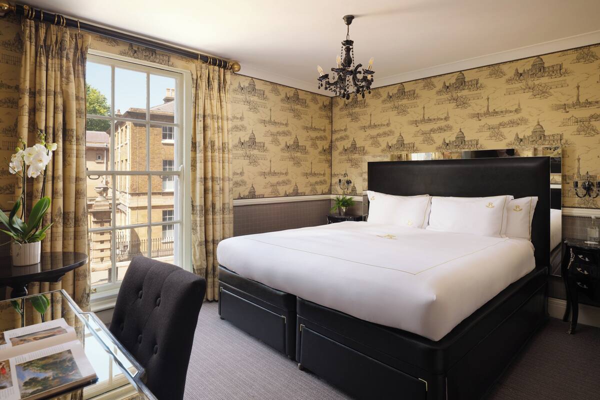 A neatly arranged hotel room with a large bed, a glass table with a chair, a potted plant, and a view of buildings outside the window.
