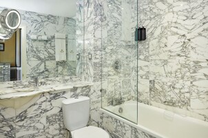 Ein Badezimmer mit Marmorwand, einer gläsernen Duschkabine, einer weißen Toilette und einem Waschbecken mit Spiegel darüber.