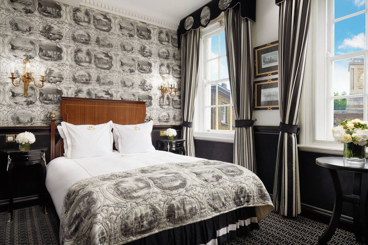 A hotel room with a large bed, a nightstand with a vase of flowers, a wall with a patterned wallpaper, and a window with curtains.