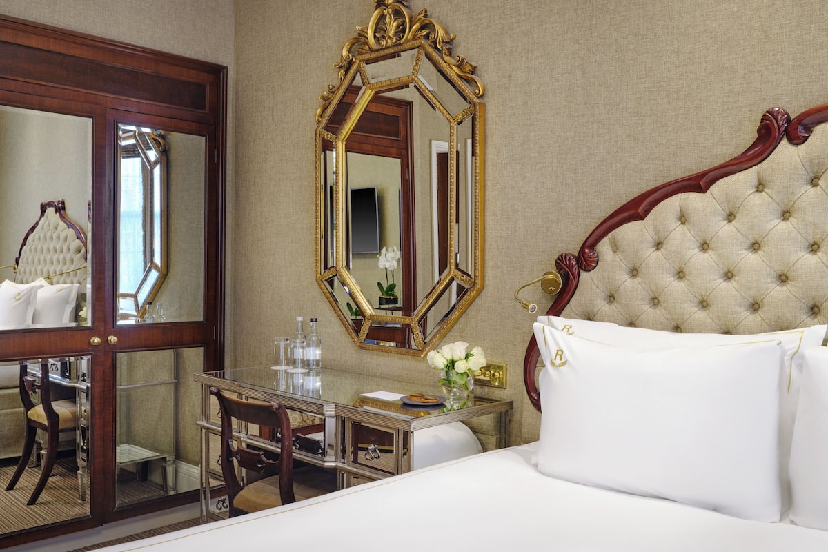 A hotel room with a large ornate mirror, a tufted headboard, a wooden desk with a chair, and a bedside table with a vase of flowers.