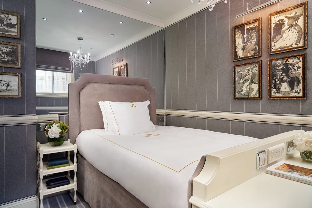 A neatly arranged bedroom with a large bed, a bedside table with books, a vase with flowers, and framed pictures on the wall.
