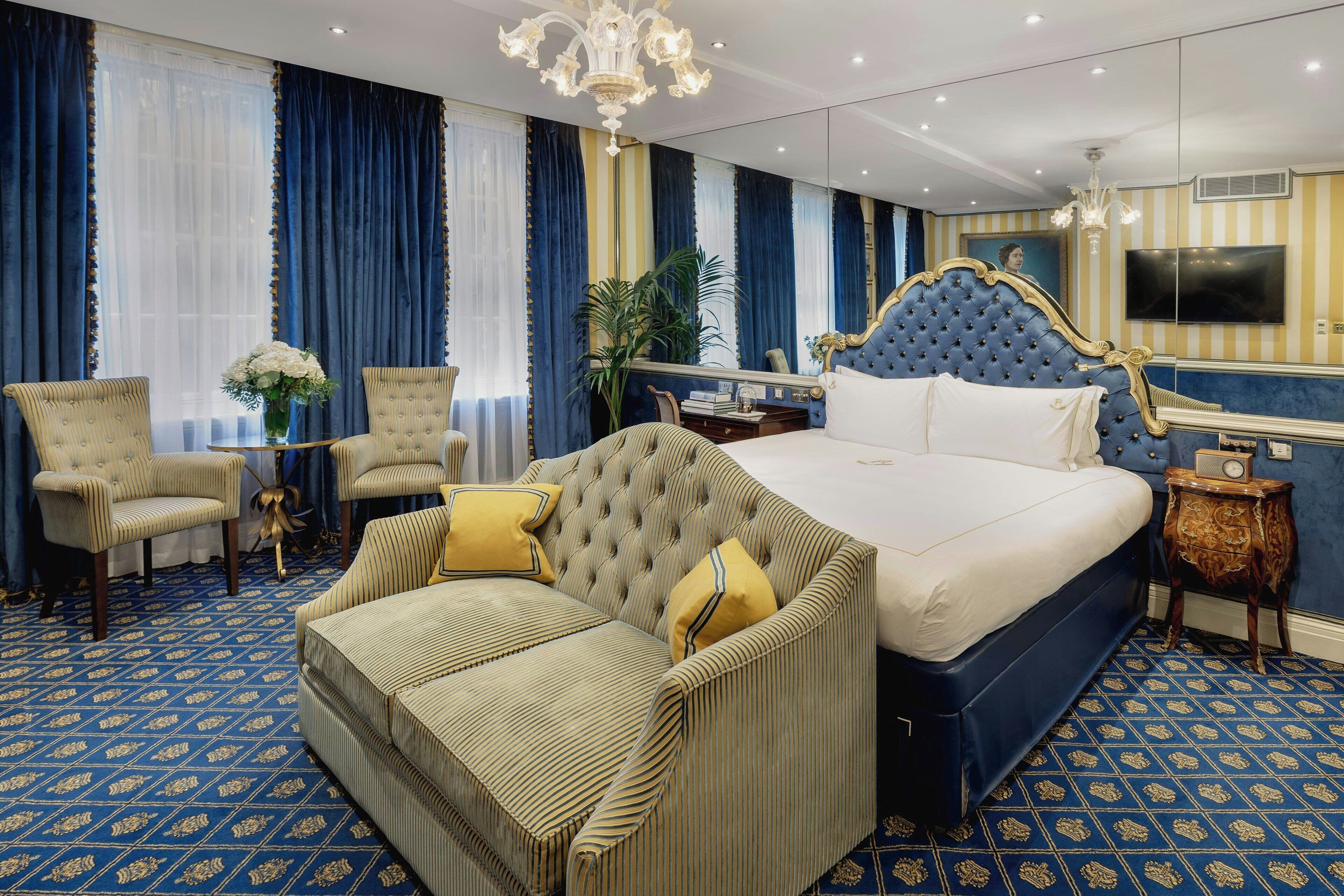 A hotel room with a large bed, a tufted headboard, a sofa, two armchairs, a small table with a vase of flowers, and a large mirror.