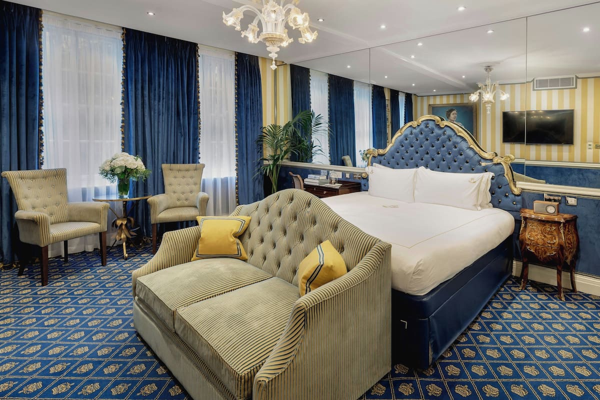 A hotel room with a large bed, a tufted headboard, a sofa, two armchairs, a small table with a vase of flowers, and a large mirror.