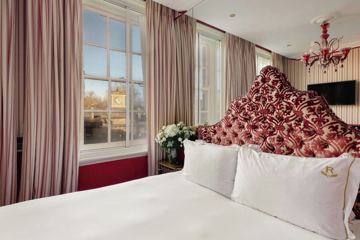 Habitación de hotel con una cama grande, un televisor montado en la pared y vista a una torre del reloj por la ventana.