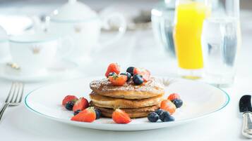 A breakfast setting with pancakes topped with strawberries and blueberries, a glass of orange juice, and a teapot in the background.