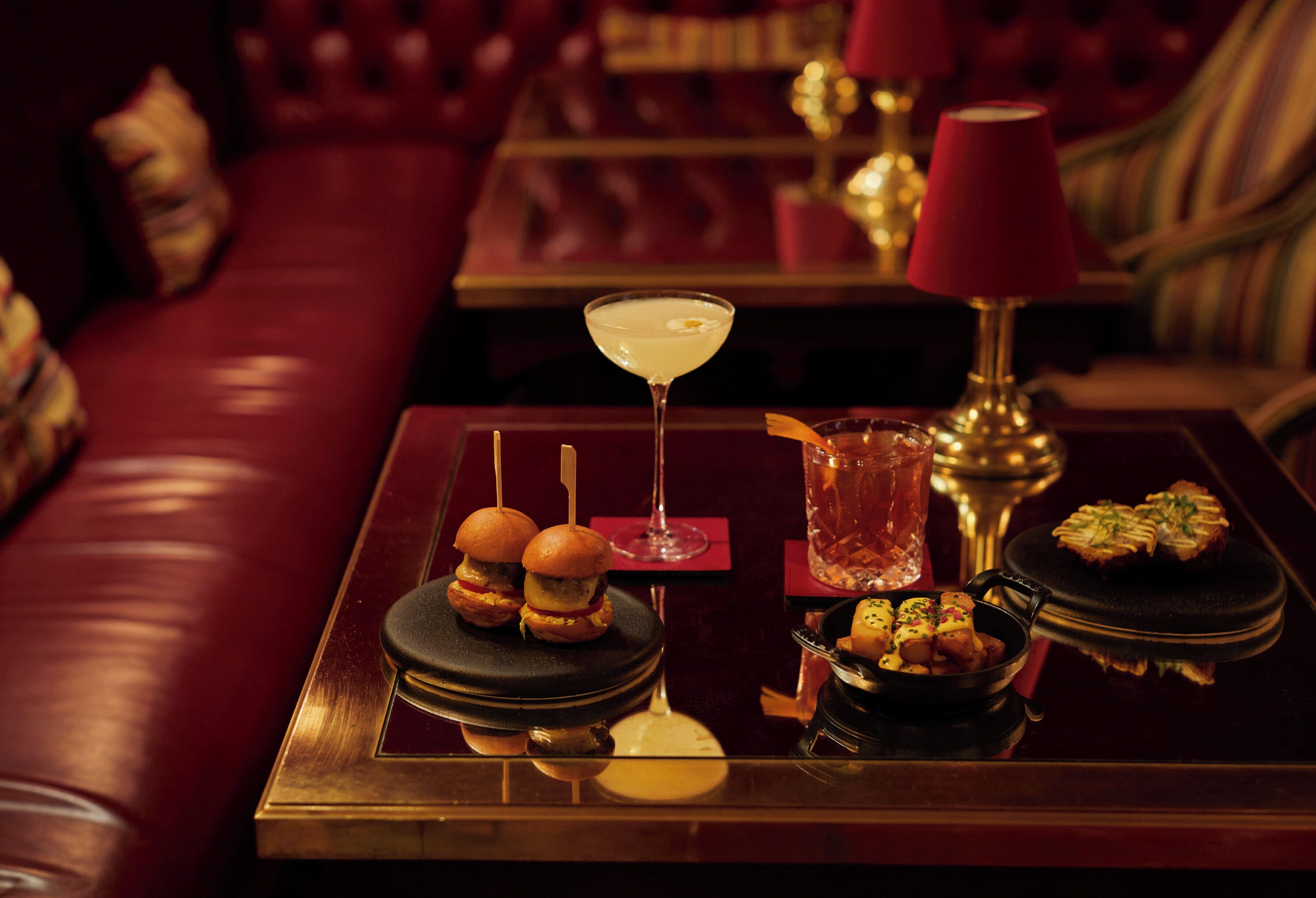 Una mesa de comedor con una copa de martini, dos hamburguesas, un vaso de agua y un pequeño plato de aperitivos.
