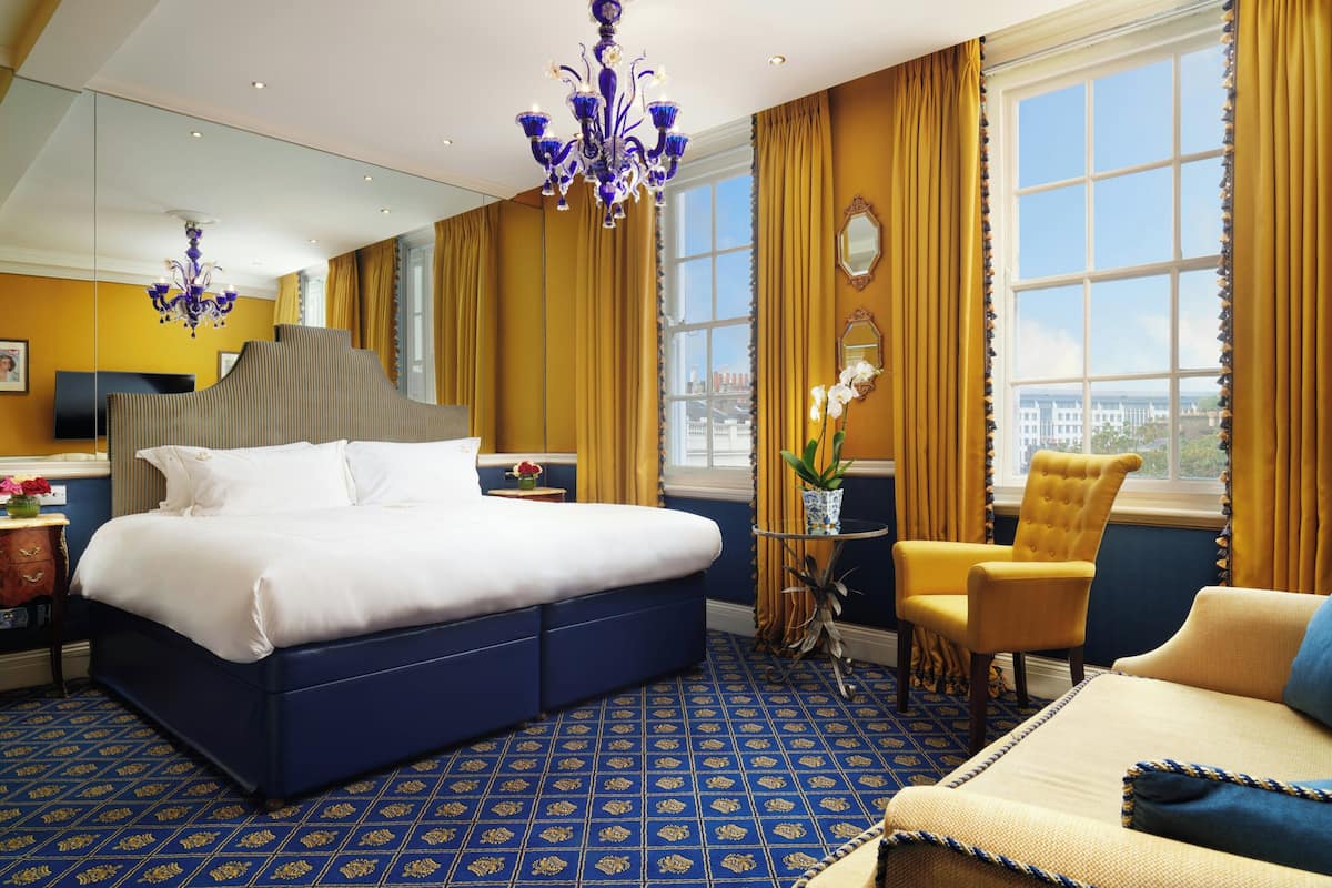 A hotel room with a large bed, a sitting area with a chair, a small table with a vase, and a view of the city through large windows.