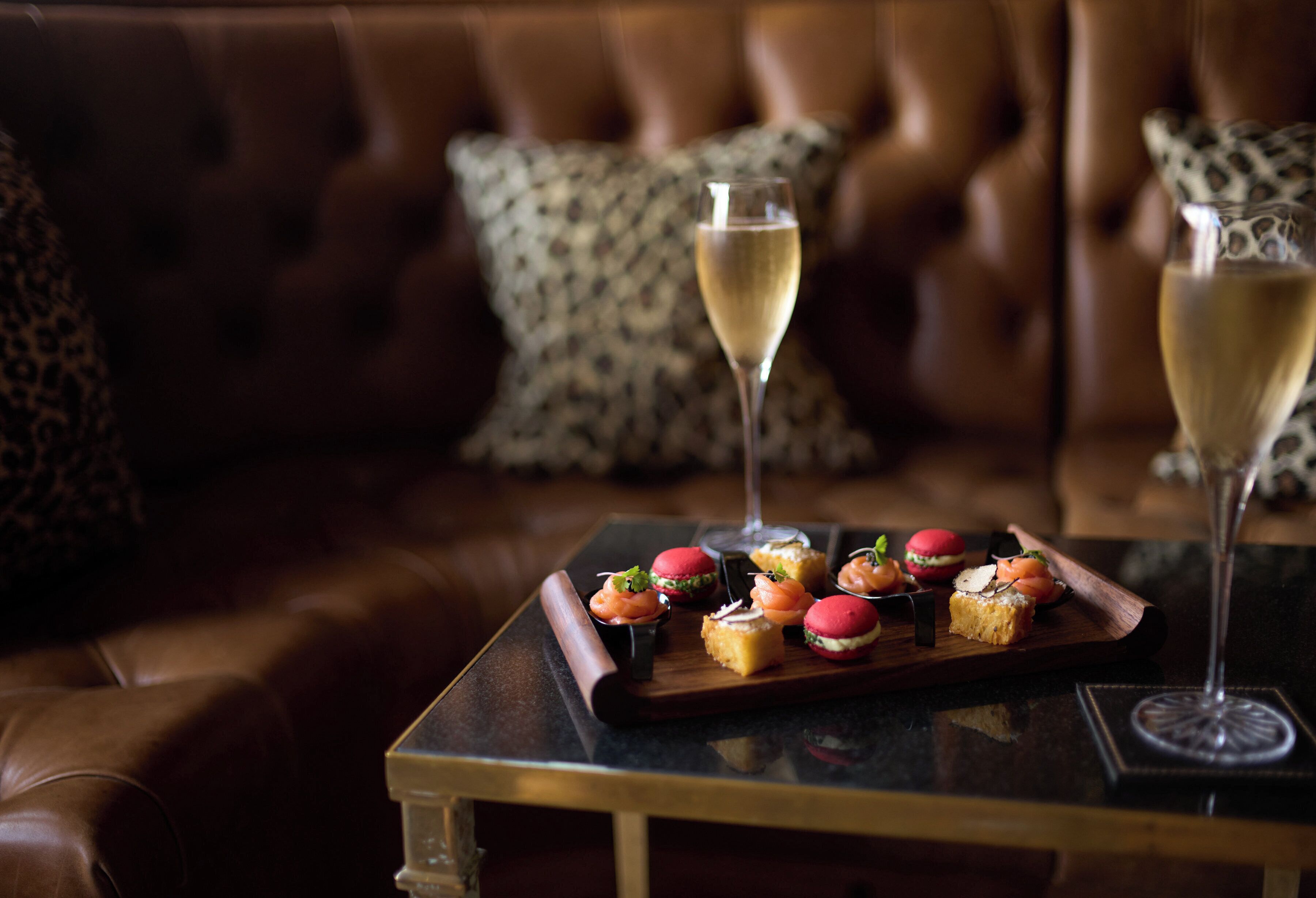 A tray of appetizers on a table with two glasses of champagne, set on a leather couch with patterned cushions.