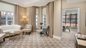 A hotel room with a patterned carpet, a sofa, a small table, a lamp, a dining area with chairs, and large windows with curtains. - Washington Mayfair Hotel (London)