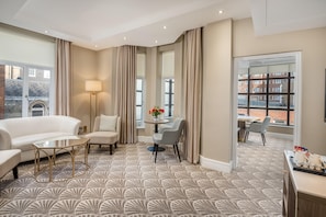 A hotel room with a patterned carpet, a sofa, a small table, a lamp, a dining area with chairs, and large windows with curtains. - Washington Mayfair Hotel (London)
